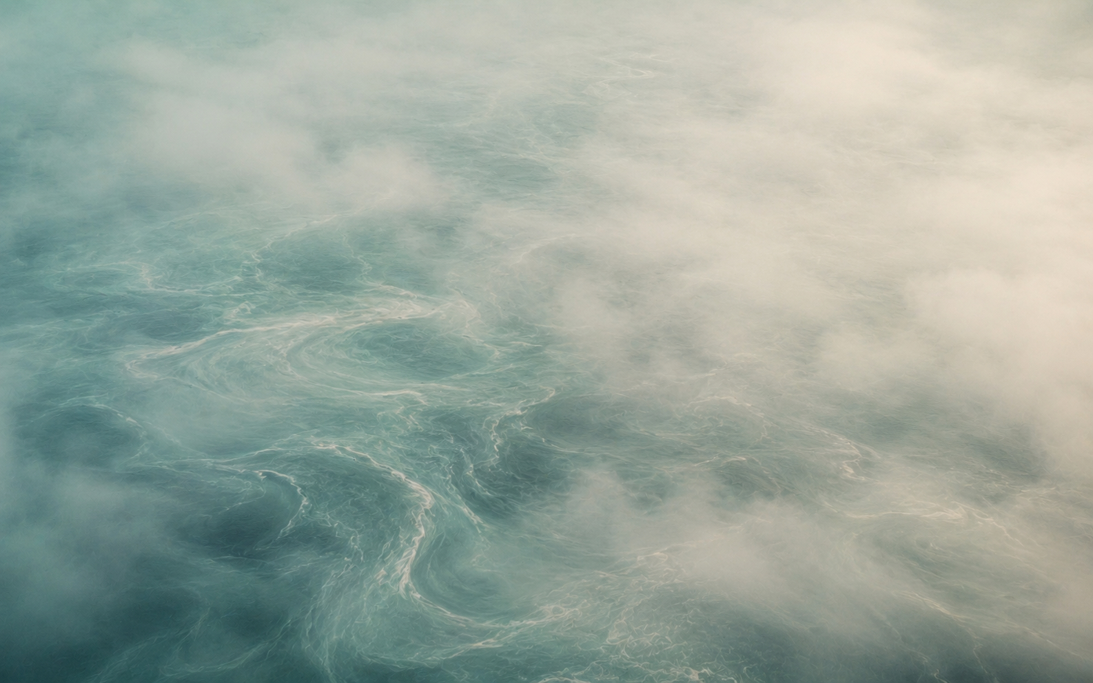 Abstract aerial image of shifting ocean currents partially obscured by fog. Soft teal and sea-glass tones move beneath layered mist, suggesting a landscape still forming rather than fully revealed.